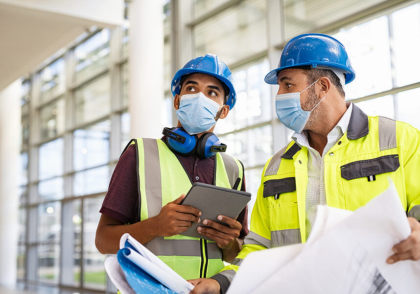 Mixed race architect and technician at construction site reviewing blueprints while wearing surgical face mask and hardhat. Team of specialists construction worker with face mask using digital tablet.